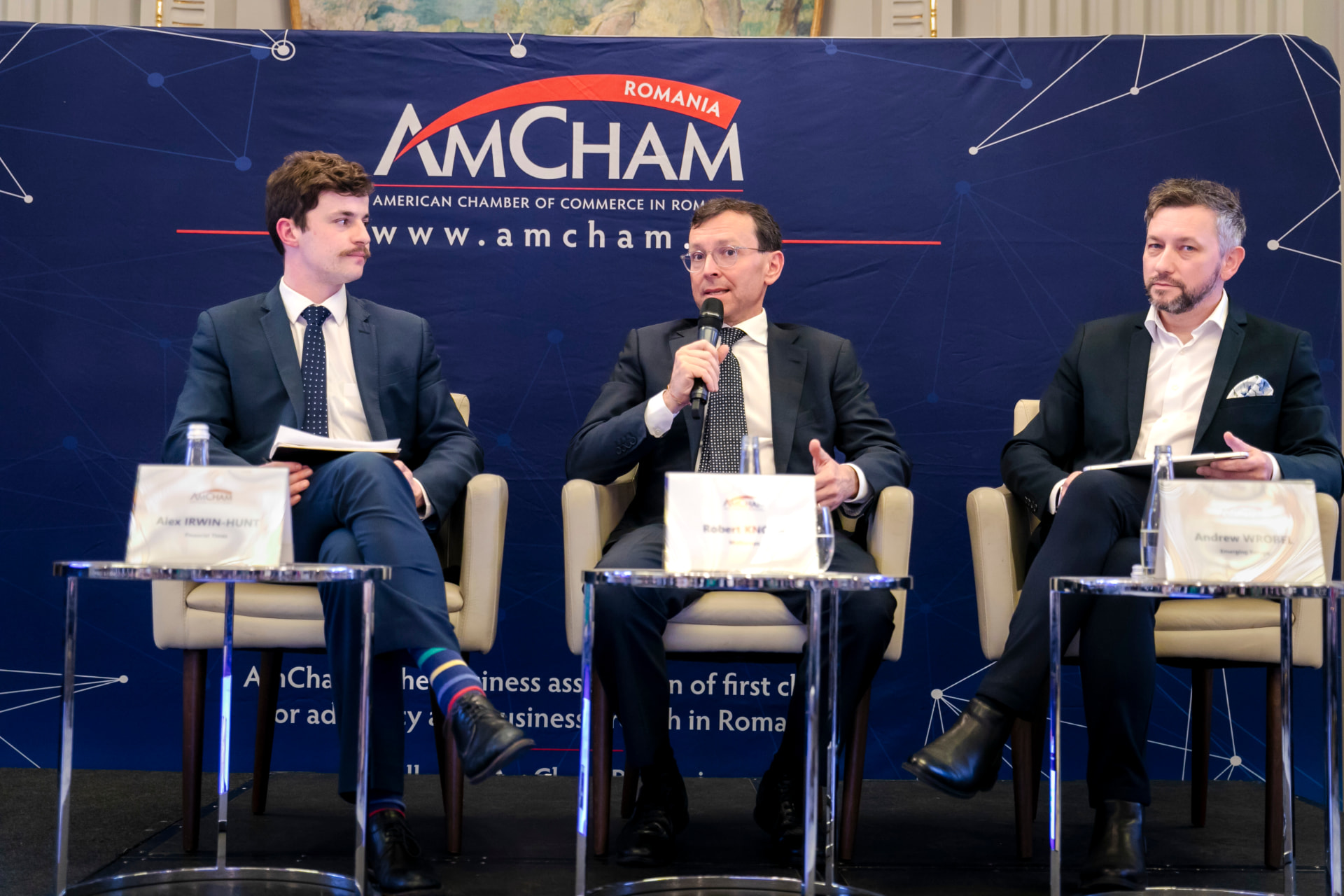 Andrew Wrobel seated on a three-person panel at the AmCham Romania Annual General Meeting in Bucharest, beside Alex Irwin-Hunt of the Financial Times, with the AmCham Romania backdrop behind the stage
