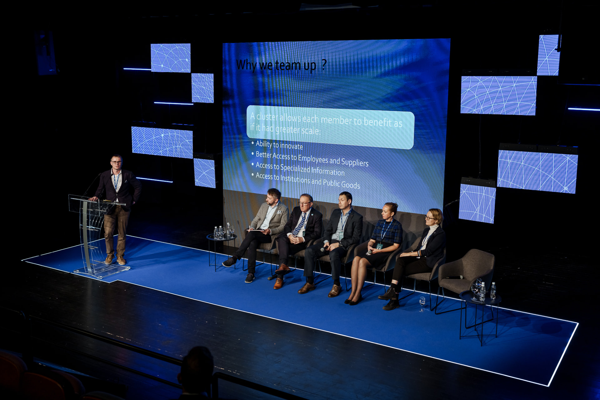 Andrew Wrobel standing at a transparent lectern at the left of a blue-carpeted stage, with five panelists seated in armchairs to his right and a screen behind them reading 'Why we team up?'