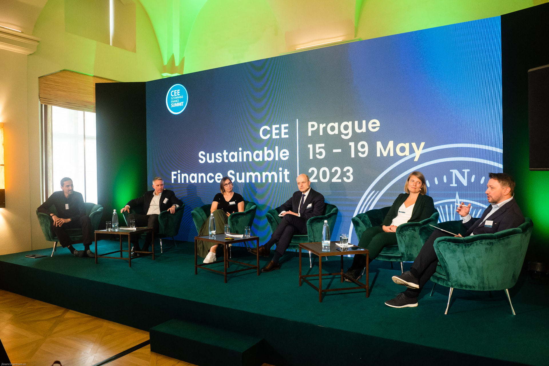 Andrew Wrobel seated among six panelists in green armchairs on a blue-lit stage at the CEE Sustainable Finance Summit in Prague, a screen behind them reading 'CEE Sustainable Finance Summit, Prague, 15–19 May 2023'