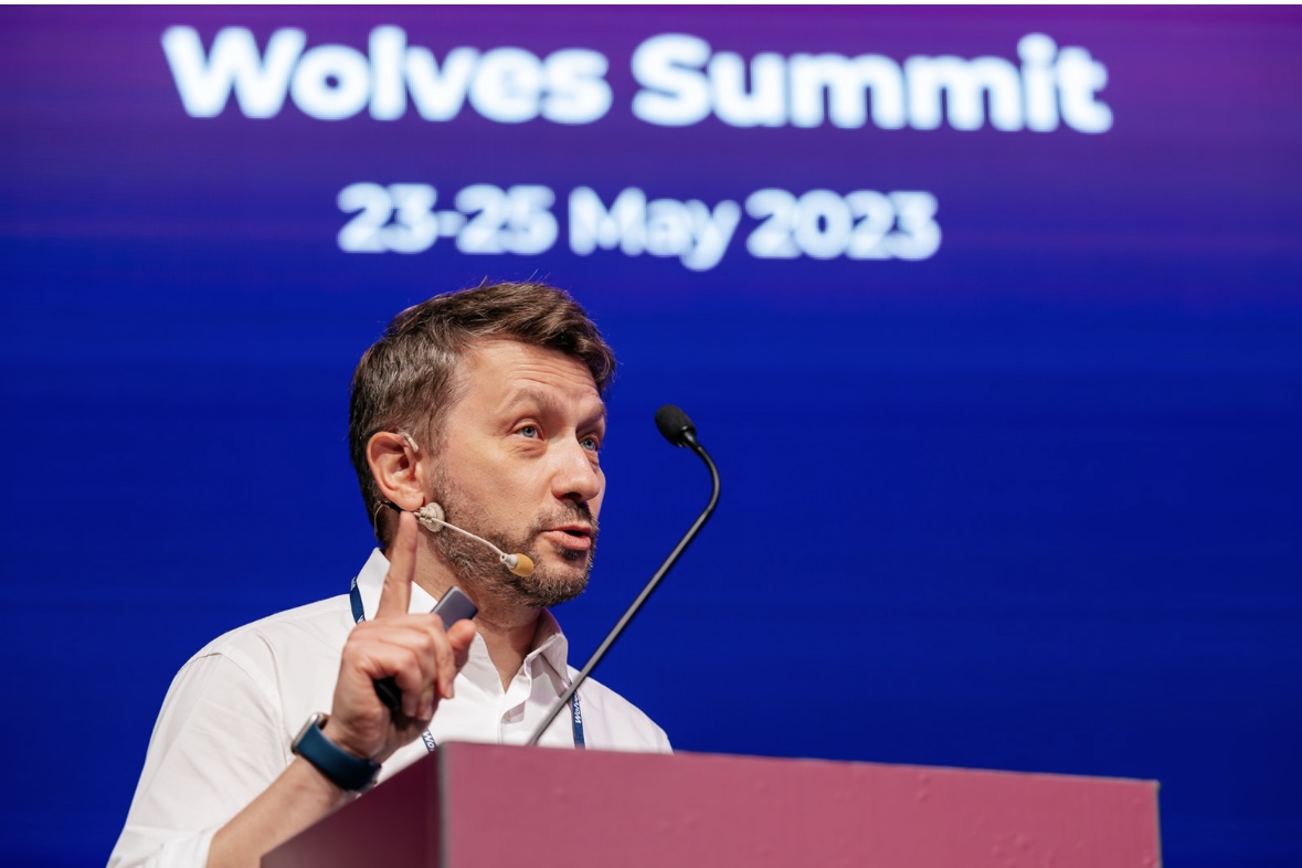 Andrew Wrobel speaking at a maroon lectern on the Wolves Summit stage in Wrocław, wearing a beige headset microphone and gesturing with one index finger raised, a blue screen behind him reading 'Wolves Summit 23-25 May 2023'