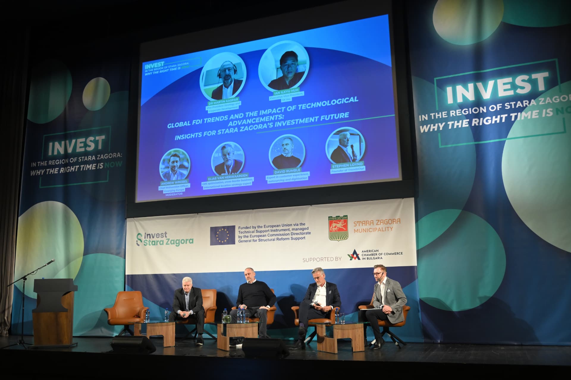 Andrew Wrobel moderating a panel at Invest in the Region of Stara Zagora, with the session screen listing the speakers behind the stage