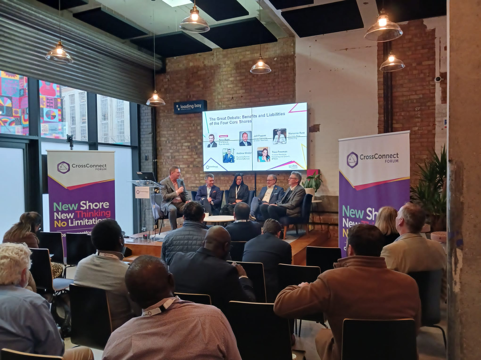 Andrew Wrobel seated with four other panelists in armchairs on a low wooden stage in a brick-walled room at the CrossConnect Forum in London, a screen behind them reading 'The Great Debate: Benefits and Liabilities of the Four Core Shores' and CrossConnect Forum banners to either side
