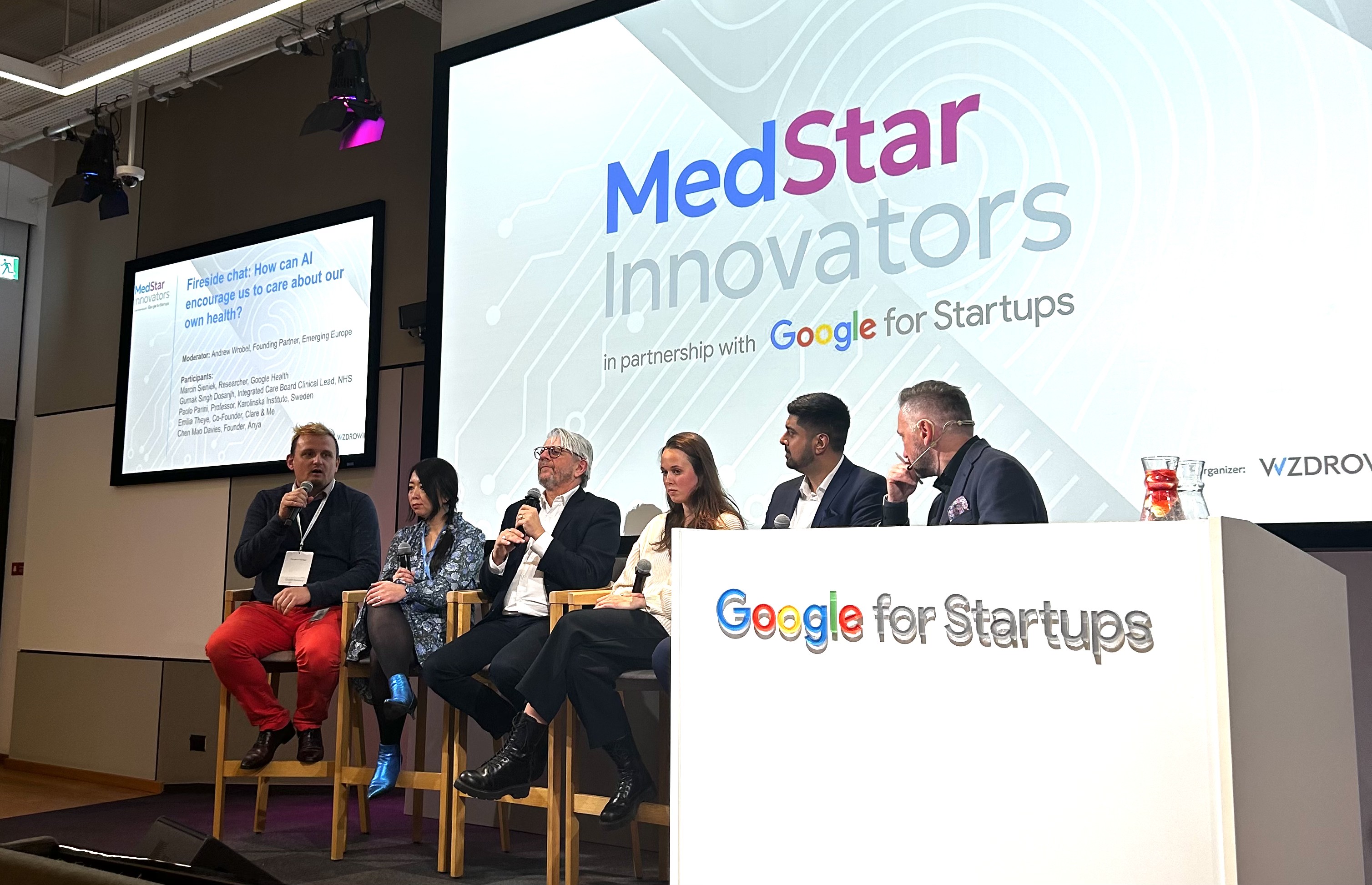 Andrew Wrobel seated at the far right of a six-person panel on tall wooden stools at MedStar Innovators in Warsaw, a screen behind the stage reading 'Fireside chat: How can AI encourage us to care about our own health?' with the MedStar Innovators backdrop in partnership with Google for Startups