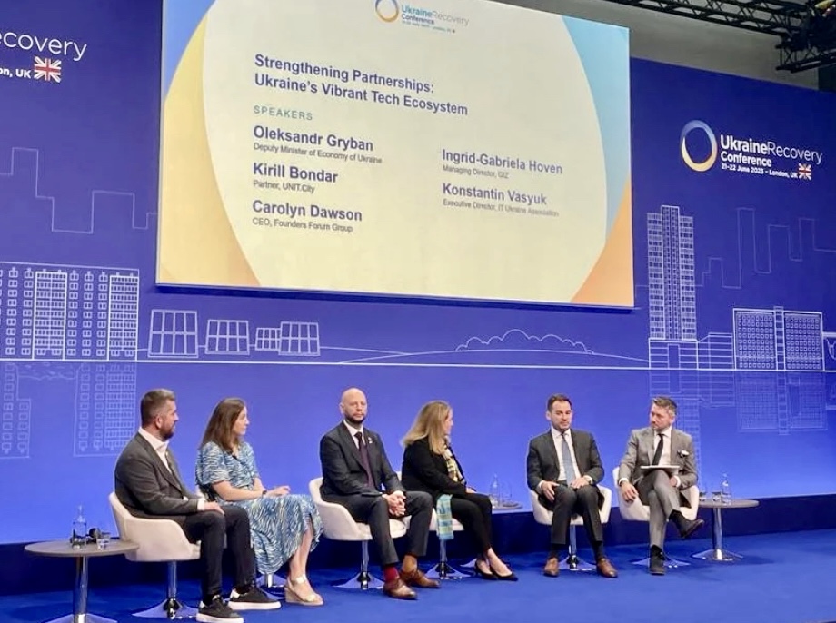 Six panelists seated in white chairs on a blue-carpeted stage at the Ukraine Recovery Conference in London, a screen above them reading 'Strengthening Partnerships: Ukraine's Vibrant Tech Ecosystem' and the conference backdrop reading 'Ukraine Recovery Conference, 21-22 June 2023 - London, UK'
