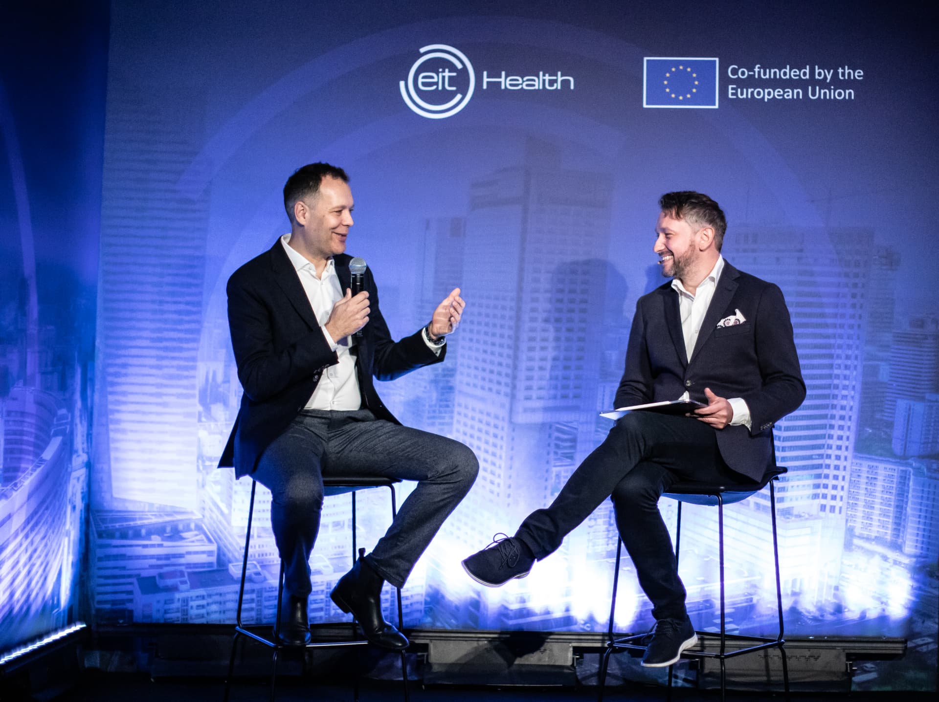 Andrew Wrobel seated on a tall stool at the right, holding notes and smiling at the man beside him who is speaking into a handheld microphone, on stage at the EIT Health InnoStars Networking Dinner in Warsaw, with the EIT Health logo and European Union flag on a blue cityscape backdrop