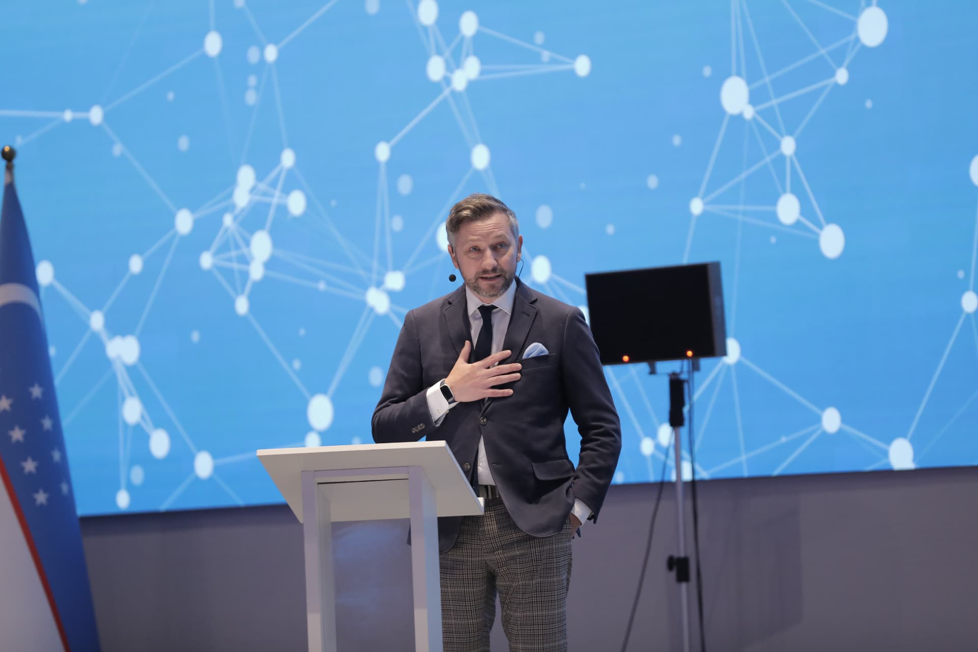 Andrew Wrobel at a lectern with his hand over his chest, delivering a keynote at the Uzbekistan Outsourcing Forum in Tashkent, with the Uzbekistan flag at the left and a blue network-graphic screen behind the stage