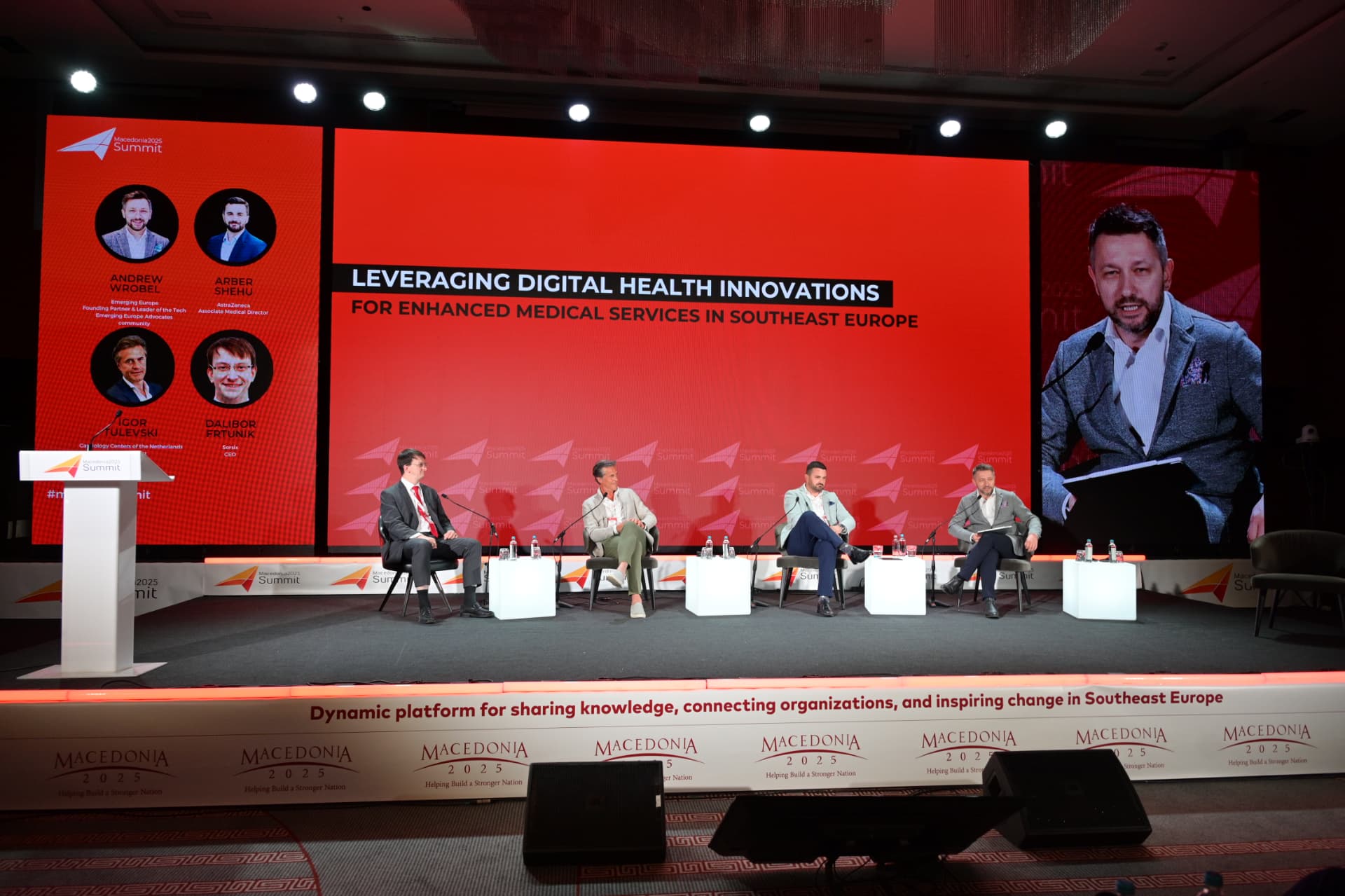 Andrew Wrobel moderating a panel from the right-hand armchair on stage at the Macedonia 2025 Summit in Skopje, holding notes and a microphone, with three fellow panelists seated to his left and a screen behind the stage reading 'Leveraging Digital Health Innovations for Enhanced Medical Services in Southeast Europe'